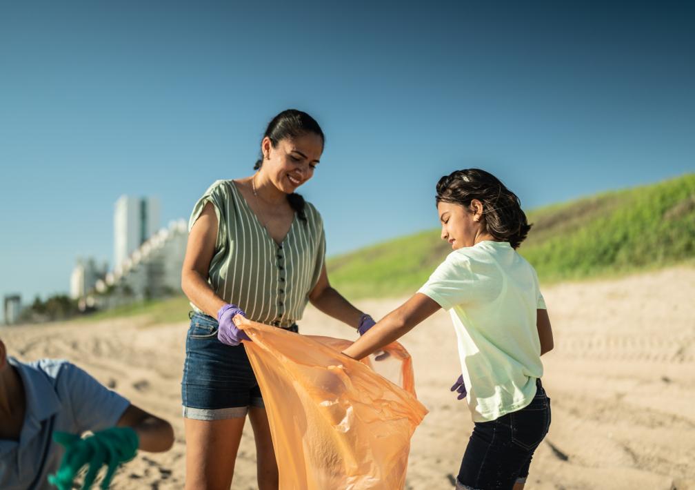 A woman holding a trash bag and a child is picking up debris from the beach.