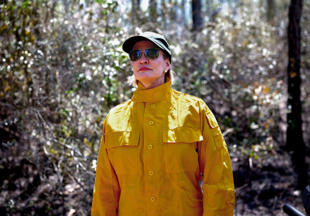 Commissioner Dawn Buckingham, M.D. wearing a raincoat, sunglasses and hat standing in front trees.