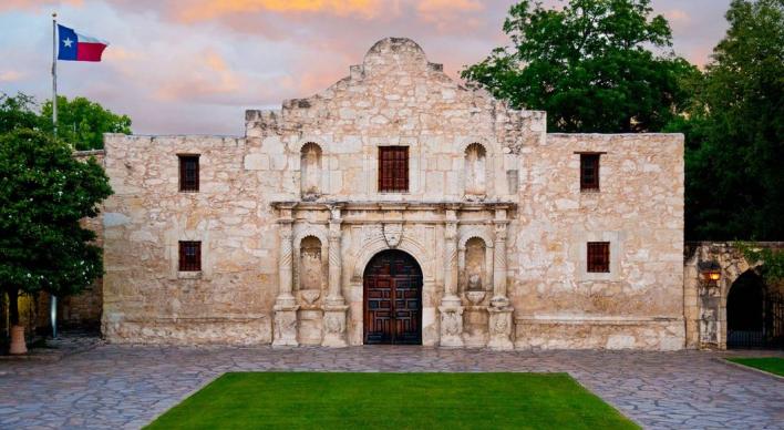 The front of The Alamo with a Texas flag, trees and sunset clouds.