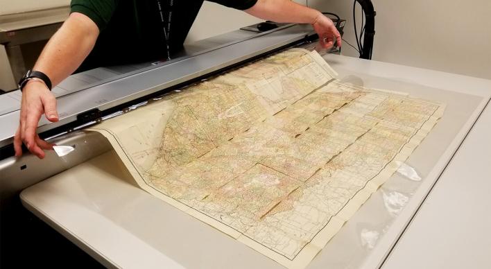 A scan lab employee feeding a original map through a large image scanner.