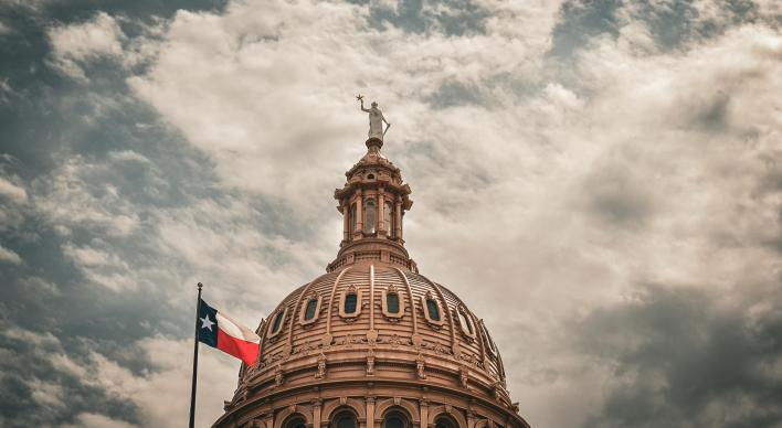 Looking up at the dome of the Texas Capitol building with Texas flag and scattered clouds in sky.