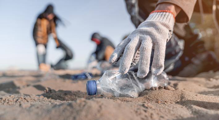 Close up of a hand picking up a plastic bottle on the beach with other volunteers in the background.